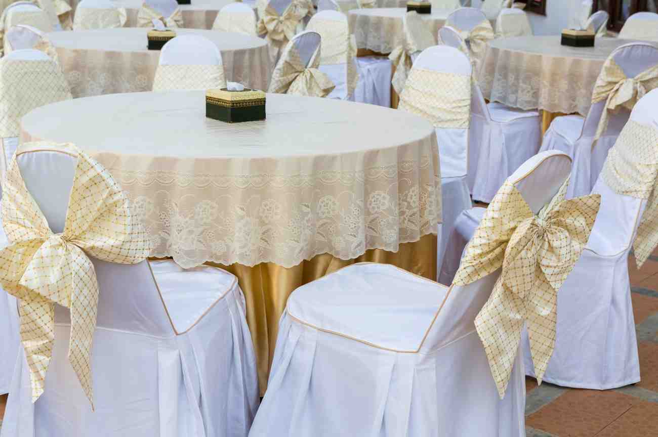 Decorated event space with round tables and chairs covered in white tablecloths and chair covers, featuring gold accents.