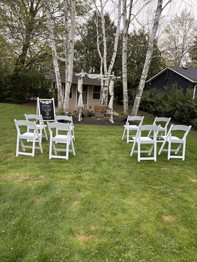 White chairs arranged in a circle on a grassy area with trees and a building in the background.