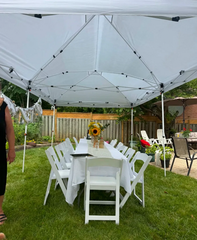 A white pop-up canopy tent set up on a green lawn in a backyard, with a long white table and white folding chairs underneath.